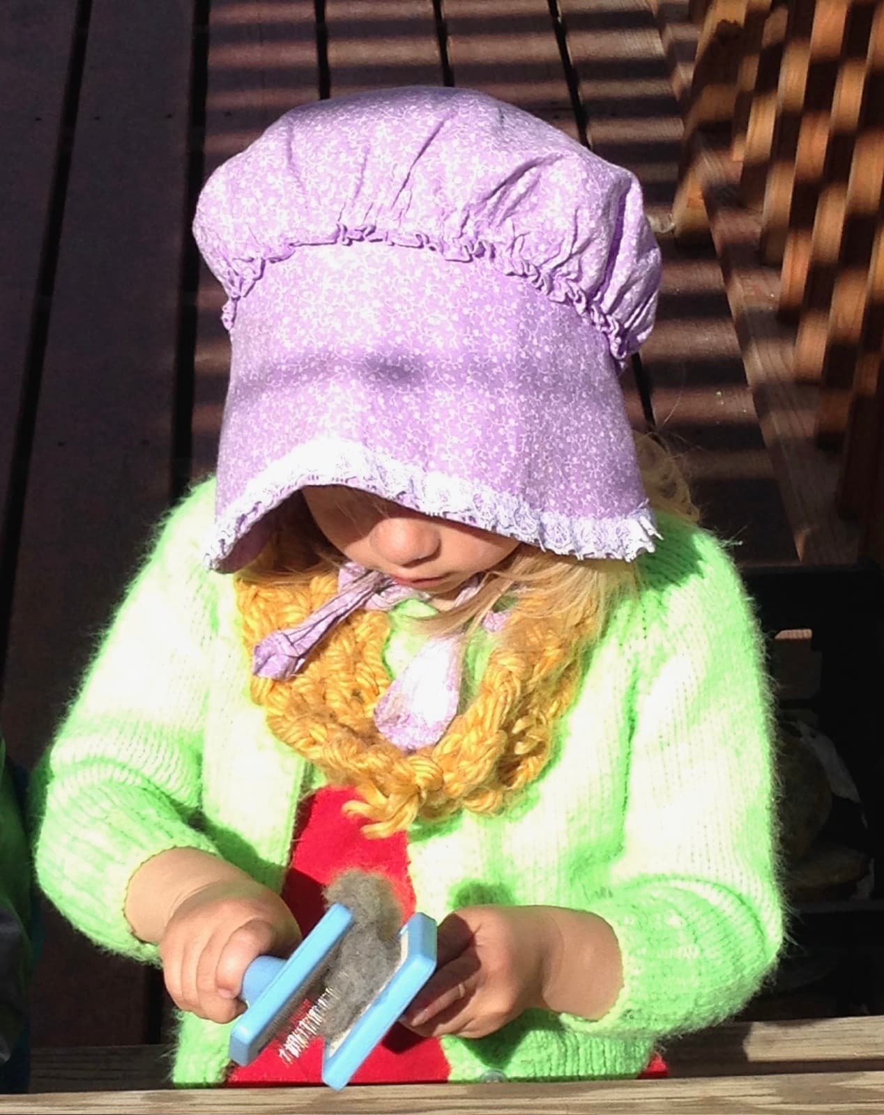 A child in a purple bonnet working a tuft of grey wool on a hand carder