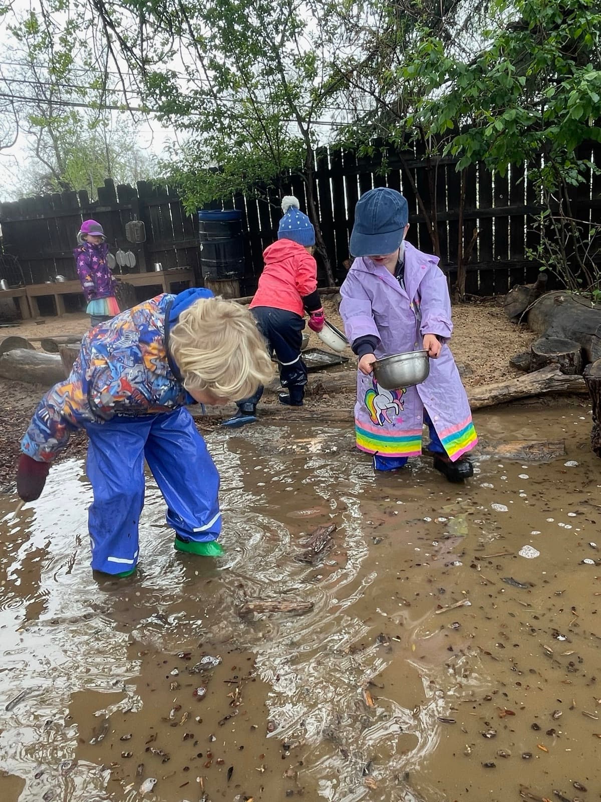 Children in rain suits and boots wading through a muddy puddle on a grey morning