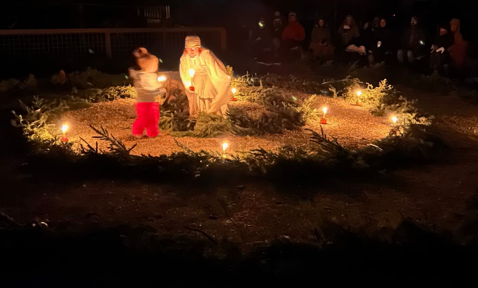 A child walking the candlelit evergreen spiral at night, reaching the light-bearer at its heart